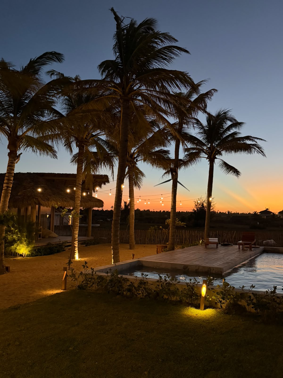 Piscina ao entardecer com coqueiros, luzes e turbina eólica ao fundo